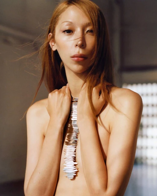 Striking close-up of a bare-chested young woman with light brown hair and pale skin, wearing a statement necklace, posing in an indoor setting.