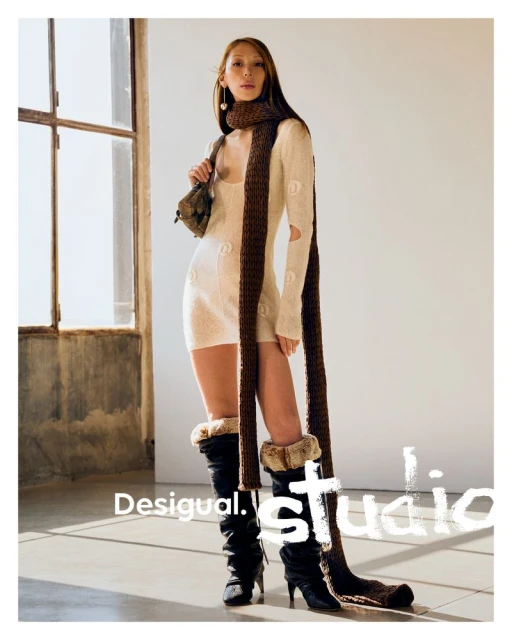 Fashion shot of a female model wearing a dress, scarf and boots in a light studio setting with sunlight streaming through a window.