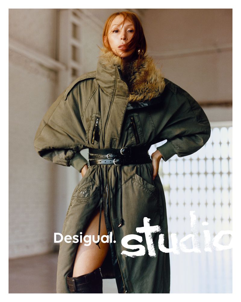 A woman in an olive-green parka-style jacket with a fur collar and a thick black belt poses indoors, with visible brand logos.