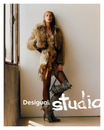 Model Issa Lish in a patterned dress and fur jacket holding a handbag against a plain wall for Desigual Studio FW25.