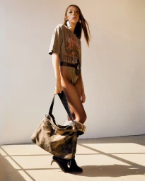 Model Issa Lish wearing a graphic t-shirt and holding a large shoulder bag against a plain wall for Desigual Studio FW25.