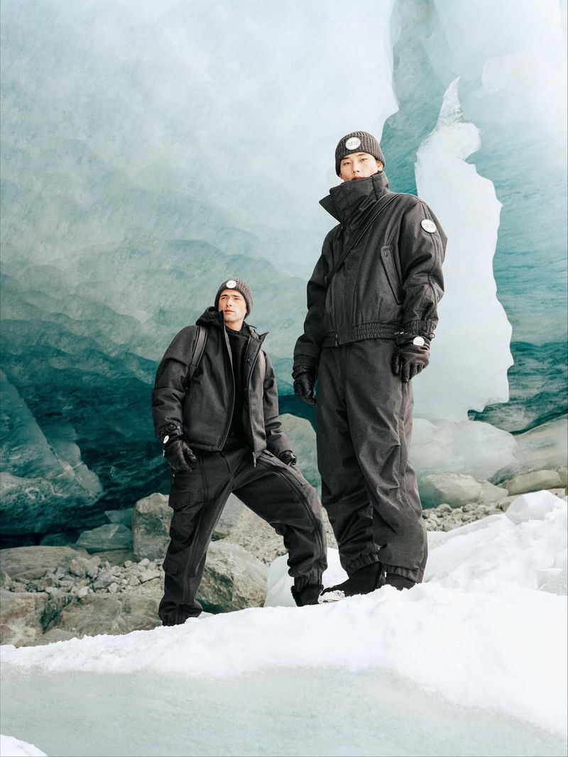 Two adult men in warm clothing stand on ice against a glacial backdrop. They pose for an outdoor fashion shot in cool tones.