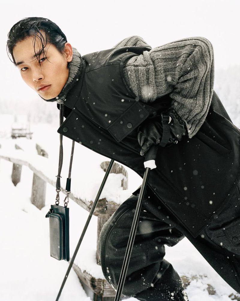 Asian man in winter apparel poses outdoors in snow with ski poles, wearing black jacket, patterned grey sweater, and black gloves.