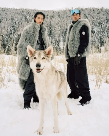 Two Asian men in fur coats pose with a dog in a snowy, wooded landscape featuring evergreen trees and wintery light.