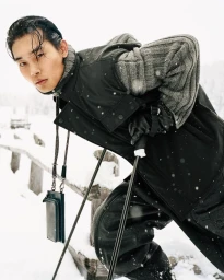 Taemin Park wearing a black utility vest and grey ribbed knit sweater holding ski poles along a snow-covered wooden fence.