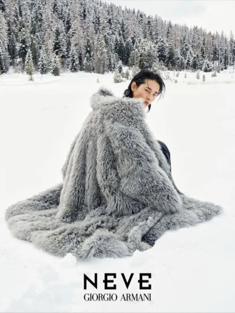 Model wearing a voluminous grey shearling coat seated in a snowy mountain landscape for Giorgio Armani Neve