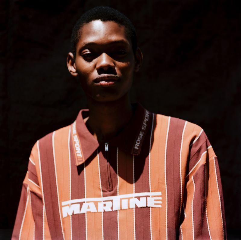Portrait of a young adult in a striped rust-colored polo shirt with 'MARINE' text, posing against a dark backdrop with soft natural lighting.