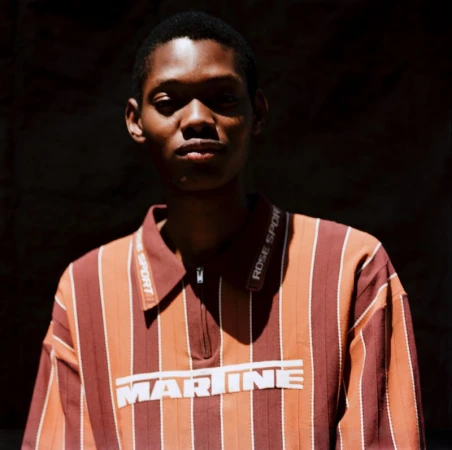Portrait of a young adult in a striped rust-colored polo shirt with 'MARINE' text, posing against a dark backdrop with soft natural lighting.