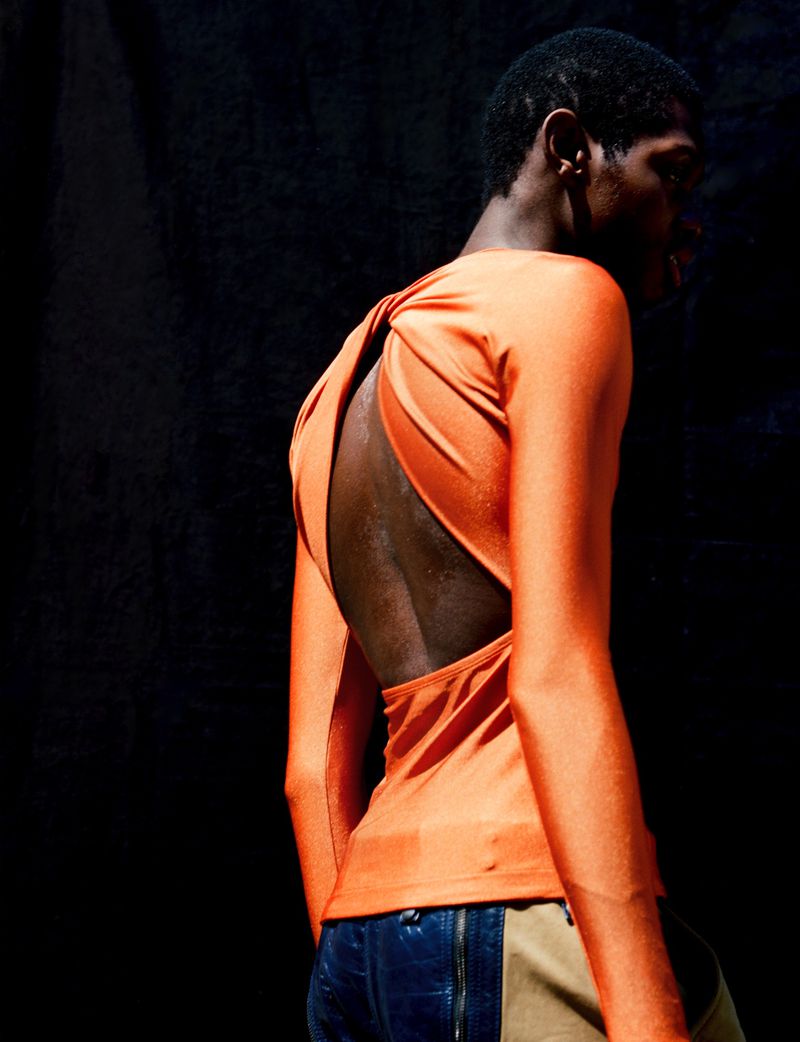 Back view of a black male model in a fitted orange top with an open back, posing in front of a dark background. Fashion portrait.
