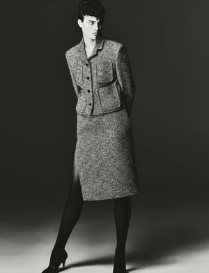 Model wearing a textured tweed jacket and matching skirt with dark tights and heels against a dark studio background.