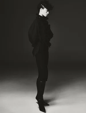 Model Niamh Battersby in dark apparel and tall boots standing against a gradient studio backdrop in a black and white editorial image