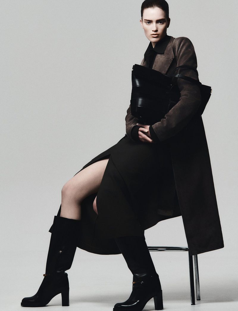 Model seated on stool wearing a dark coat and knee-high boots holding a large black bag against a grey background
