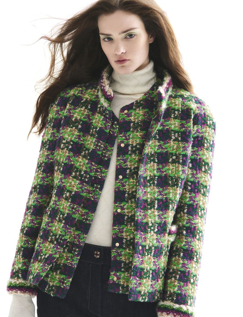 A woman poses in a green, blue, and pink plaid jacket and dark blue jeans against a bright white background in a fashion studio shot.
