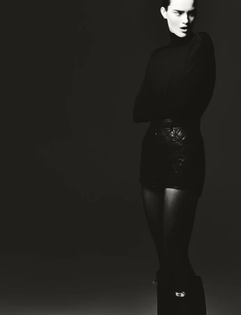 Monochromatic portrait of a woman in a turtleneck and patterned shorts with arms crossed, posing against a dark background.