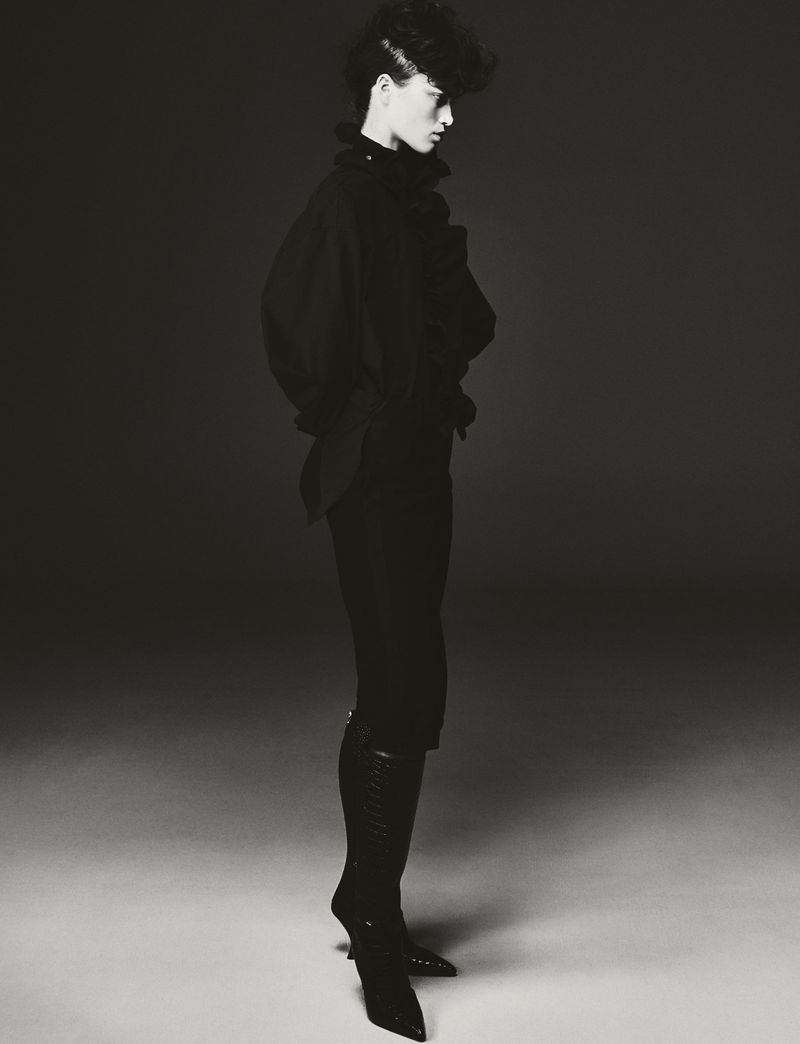 A person in an all-black outfit stands in profile, modeling high-heeled boots in a studio with dark lighting and a gradient background.
