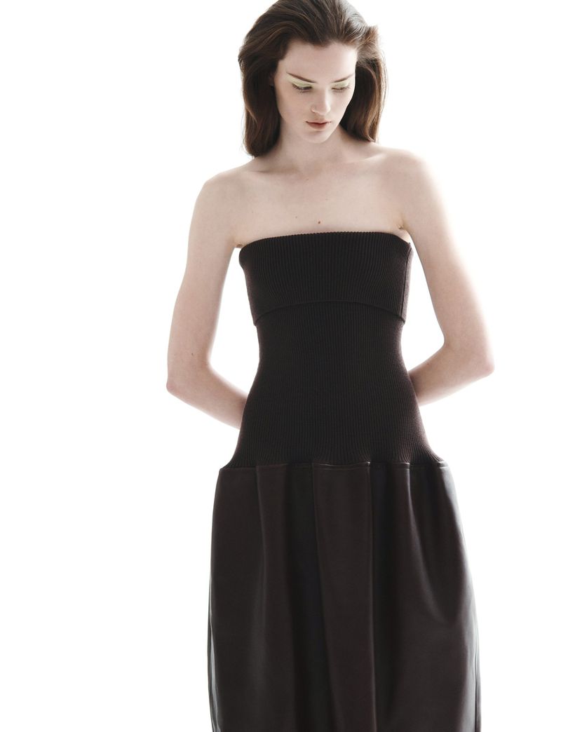 A slender female model is showcased in a studio setting, wearing a sophisticated black strapless dress with a ribbed bodice and flowing skirt.