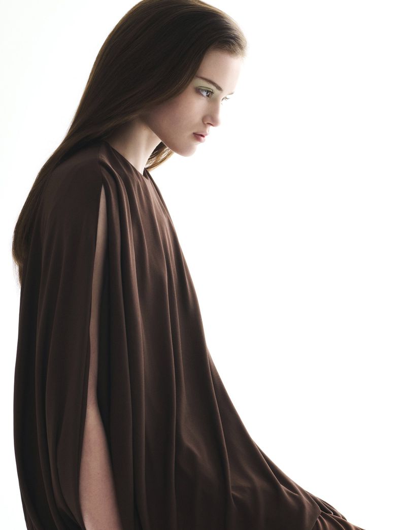 Elegant young woman in a flowing brown dress poses in a high-key studio setting. She is looking down in a side profile view.