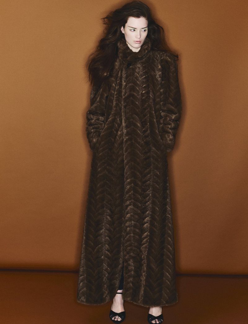 A slender woman with long brown hair is gracefully posing in a patterned floor-length brown coat and open-toe black heels in studio.