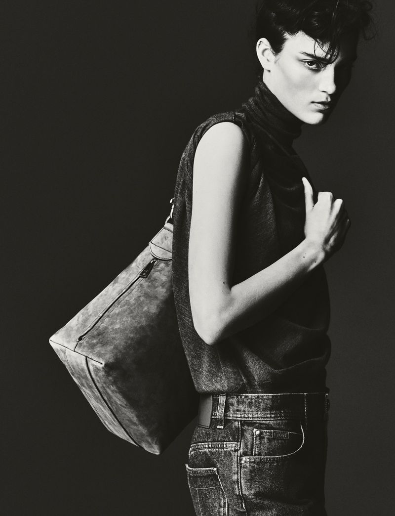 Monochrome portrait of a person with short dark hair wearing a sleeveless top and jeans, carrying a large shoulder bag.