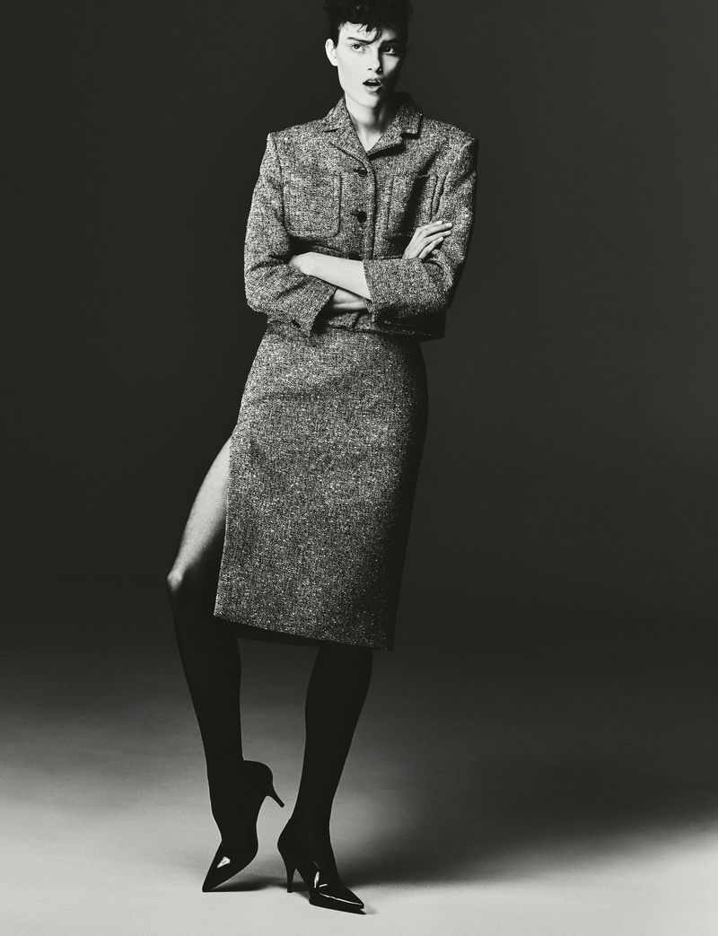 Monochrome portrait of a woman in a tweed suit with a skirt featuring a high slit, paired with black tights and high heels.