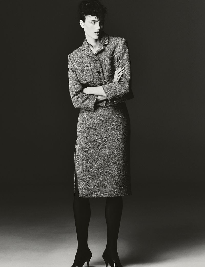 Monochrome portrait of an adult female fashion model with short dark hair posing in a tweed skirt suit with black tights and heels.