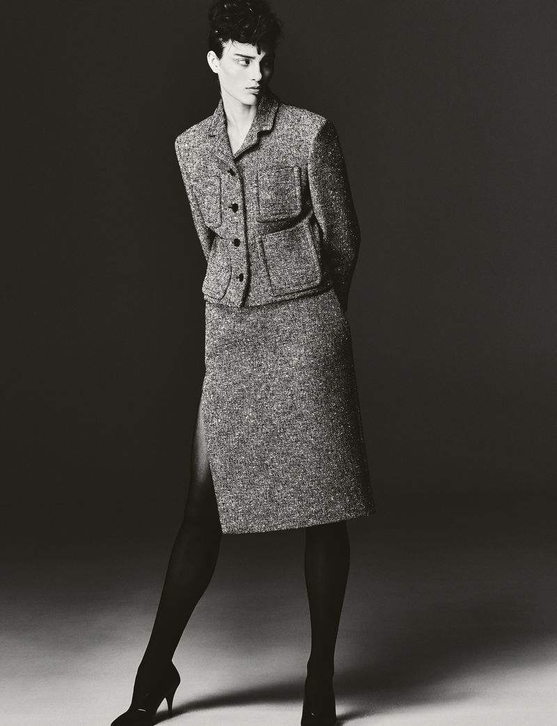 A woman in a tweed skirt suit and black tights poses in a studio with hands behind her back, standing under bright, directed lighting.