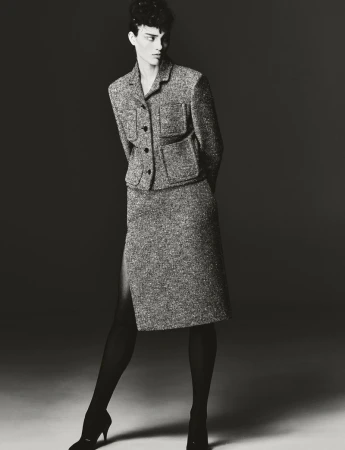 A woman in a tweed skirt suit and black tights poses in a studio with hands behind her back, standing under bright, directed lighting.