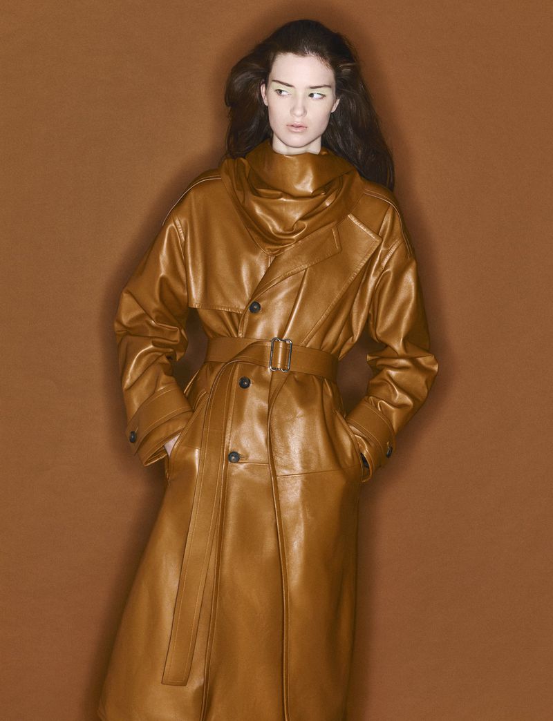 Brunette woman posing in studio wearing tan leather-look belted trench coat against a solid tan background, fashion studio shot.