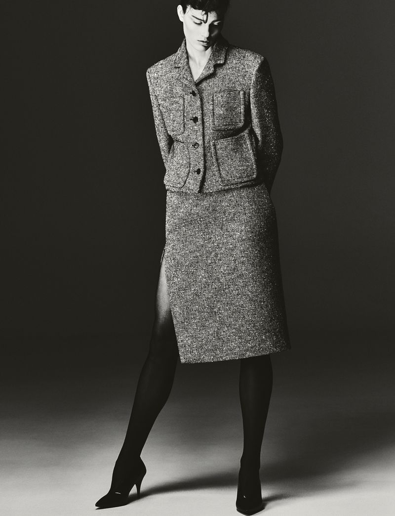 Monochrome portrait of a woman in a textured grey skirt suit, standing in a photo studio with a dark grey background, wearing black tights and heels.