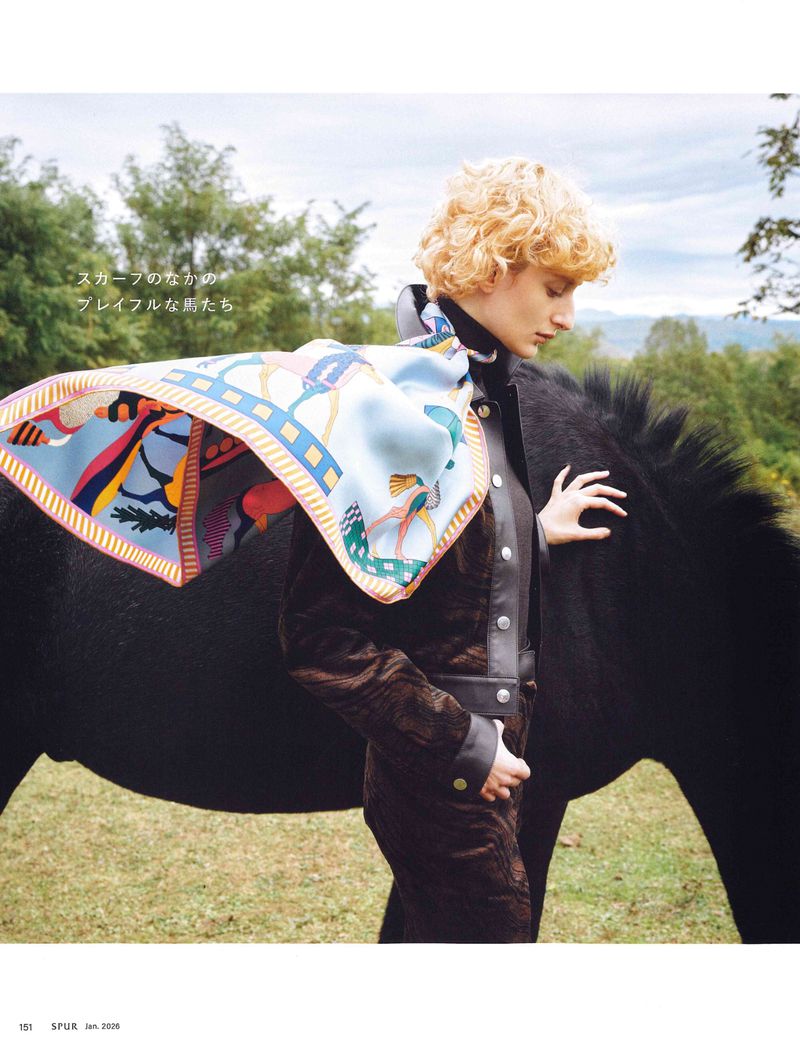 Woman in a brown jacket stands next to a black horse outdoors, wearing a patterned scarf. Equestrian fashion and rural setting.