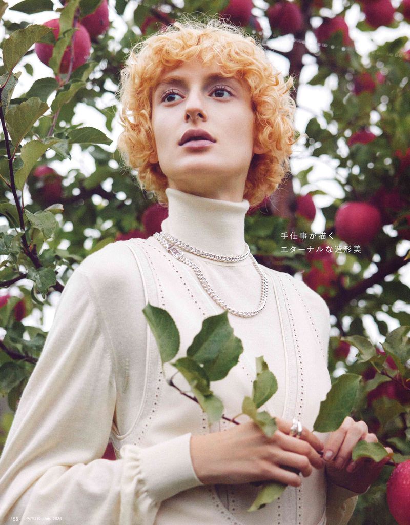 Blonde woman in white turtleneck sweater poses among apple tree branches filled with red apples, modeling in outdoor setting.