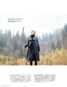 Model Alcestes Shaba holding a rustic wooden staff outdoors in a tall dry grass field with dense pine trees under an overcast sky.