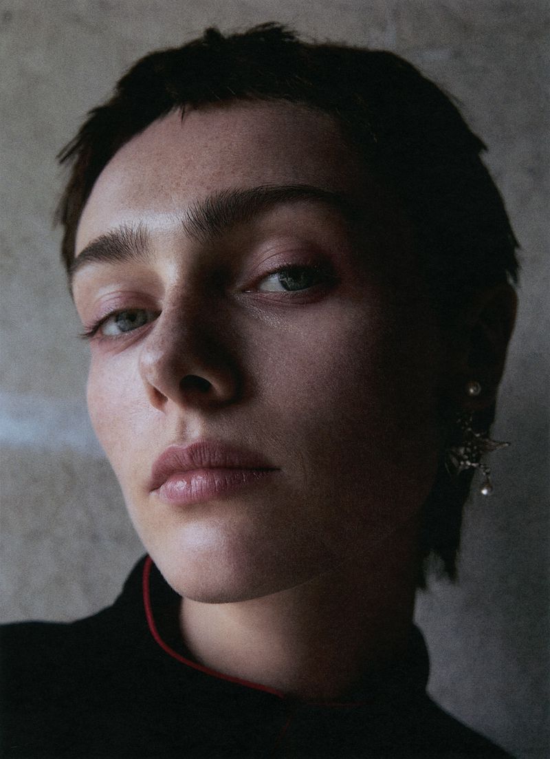 Close-up portrait of a young adult woman with short dark hair, wearing a black top with a red neckline and jeweled earring against a grey background.