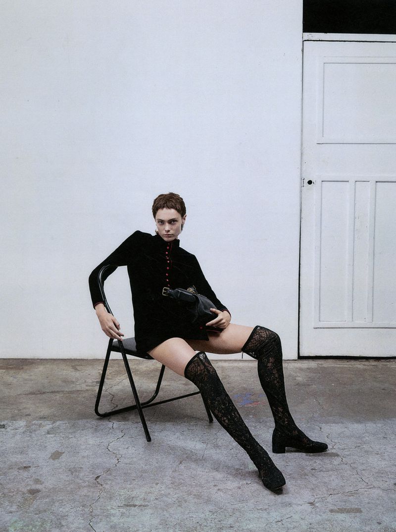A woman in a black dress and lace tights sits on a chair in a photo studio holding a purse, with a white wall and door in the background.