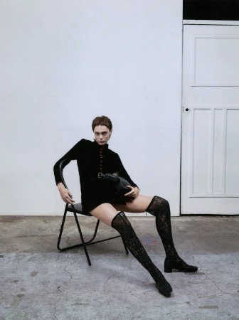 A woman in a black dress and lace tights sits on a chair in a photo studio holding a purse, with a white wall and door in the background.