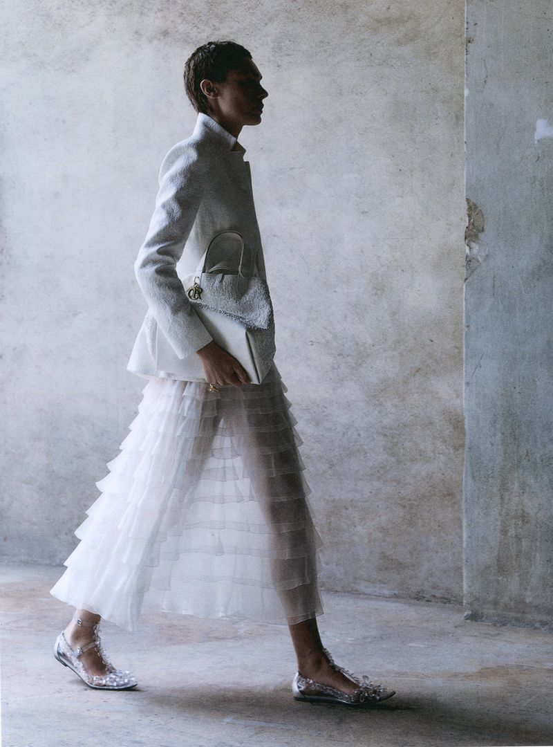 Fashion shot of a slender woman with short hair walking in a textured white blazer, layered white skirt, white purse and shiny shoes.