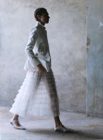 Fashion shot of a slender woman with short hair walking in a textured white blazer, layered white skirt, white purse and shiny shoes.