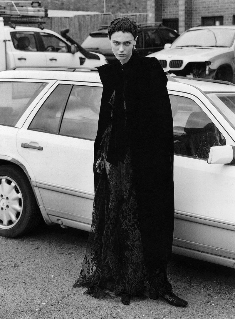 Monochrome portrait of a woman in a black cape and dress, standing on an asphalt road near parked cars and a brick building.