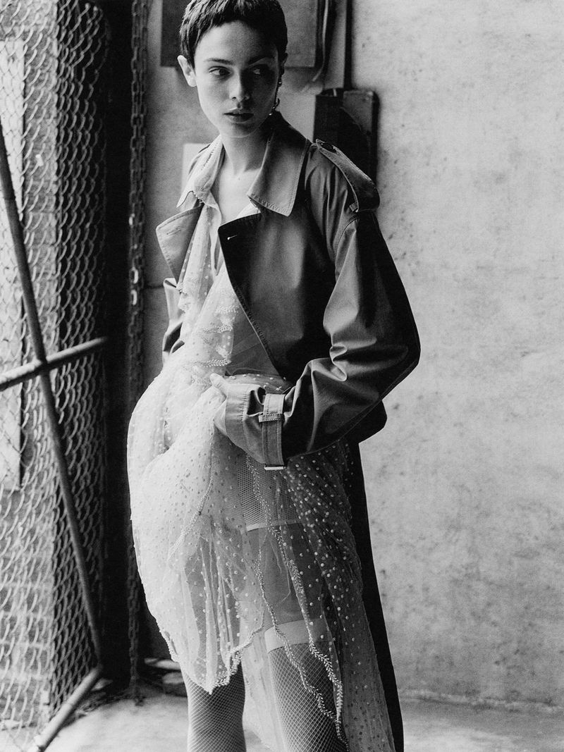 Monochrome fashion portrait of a young woman in a trench coat and layered tulle dress, with a metal gate and textured wall backdrop.
