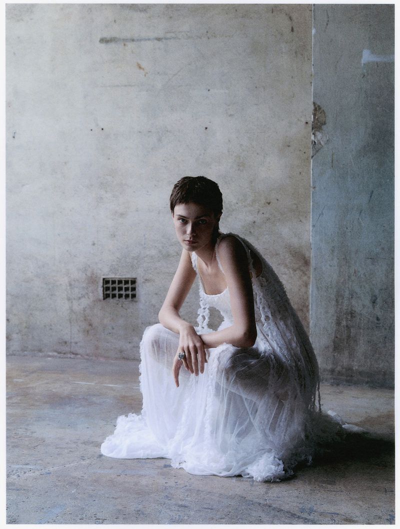 A woman in a white dress poses in a sparse room. The woman has short hair and is looking at the camera. Light is soft and even.