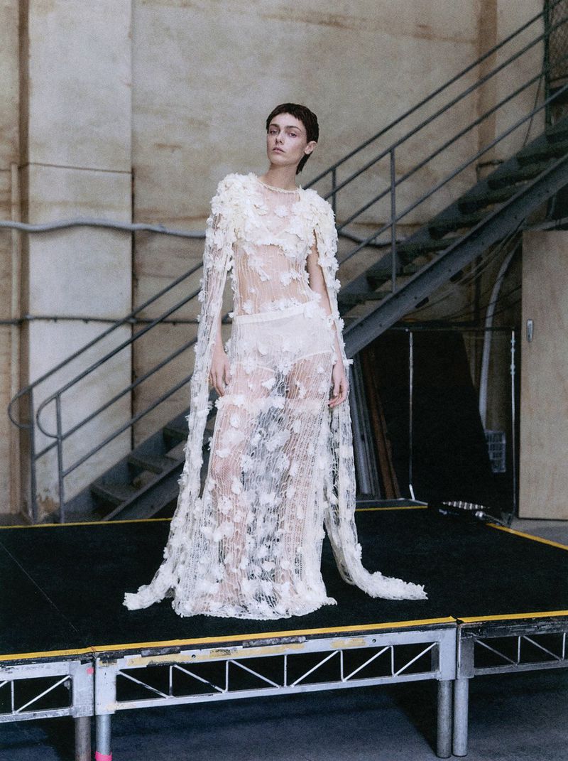 A model poses on a platform wearing a sheer white gown with floral embellishments, inside a studio with visible pipes and stairs.
