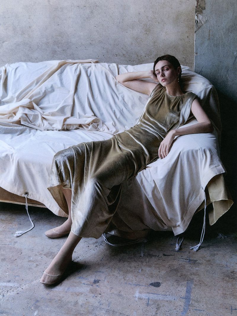 Elegant woman in a gold dress sits on a draped couch, posing in a minimalist indoor setting, showcasing fashion and style.