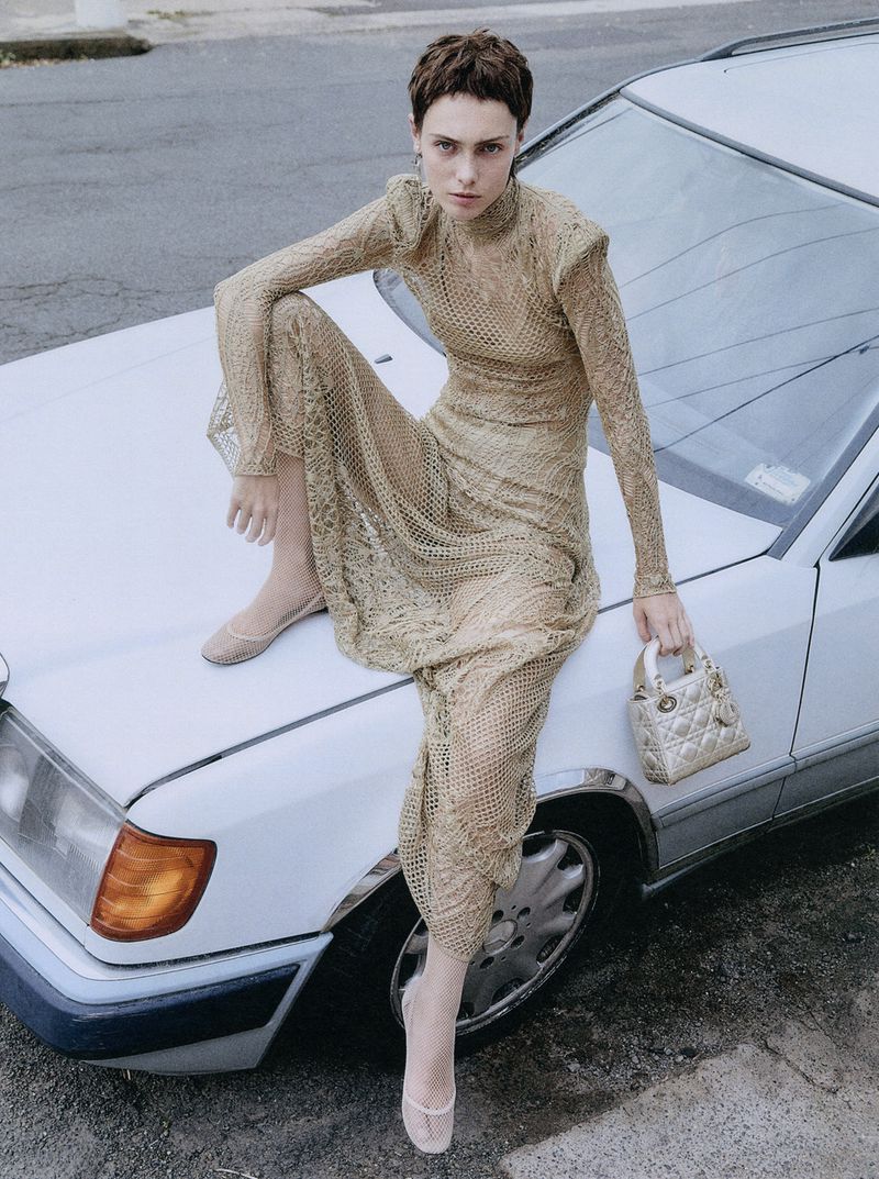 A stylish woman sits on a white car in a long, gold, see-through dress, holding a small bag in an outdoor urban setting on a sunny day.