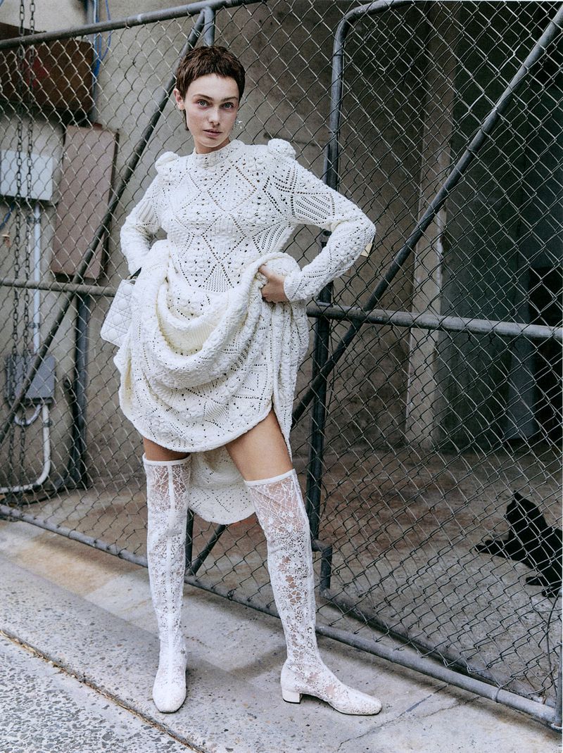 Woman with short brown hair poses outdoors in a white knit dress and lace boots, standing in front of a chain-link fence.