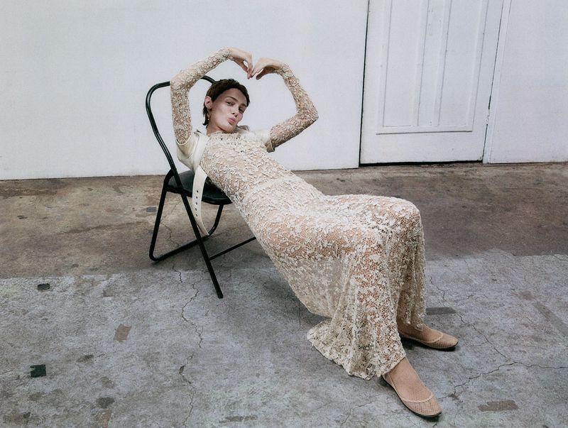 Young woman poses in a lace dress while seated on a chair in a minimalist indoor setting, casting a direct gaze toward the viewer.