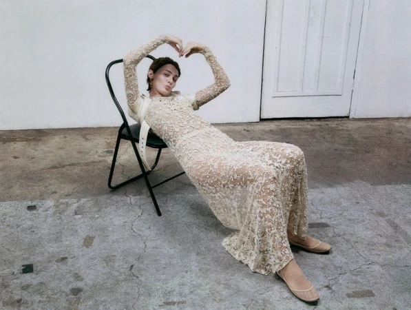 Young woman poses in a lace dress while seated on a chair in a minimalist indoor setting, casting a direct gaze toward the viewer.