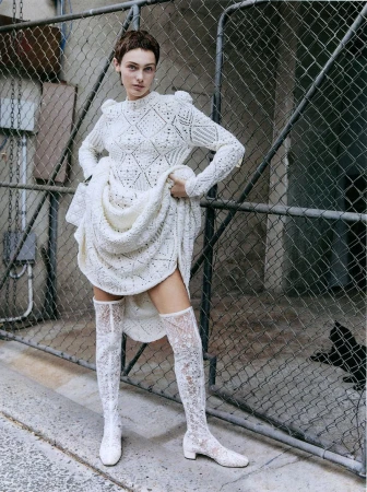 Model Sophia Roberts wearing a white patterned knit dress and matching lace boots against a chain-link fence.