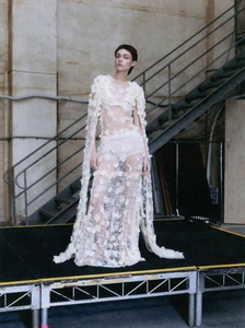 Model Sophia Roberts wearing a sheer white floral applique gown standing on a stage near metal stairs