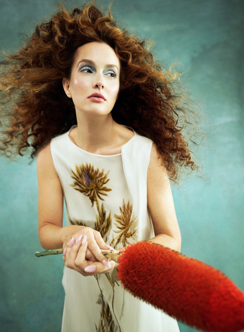 A woman with large curly auburn hair, wearing a flower-print dress, poses indoors with a red thistle-like flower. Fashion and beauty.