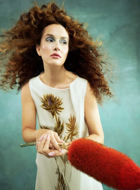 A woman with large curly auburn hair, wearing a flower-print dress, poses indoors with a red thistle-like flower. Fashion and beauty.